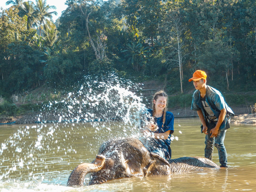 Elefant steht tief im Flusswasser und spritzt Frau und Mann an, die auf Rücken des Elefanten sitzen und stehen